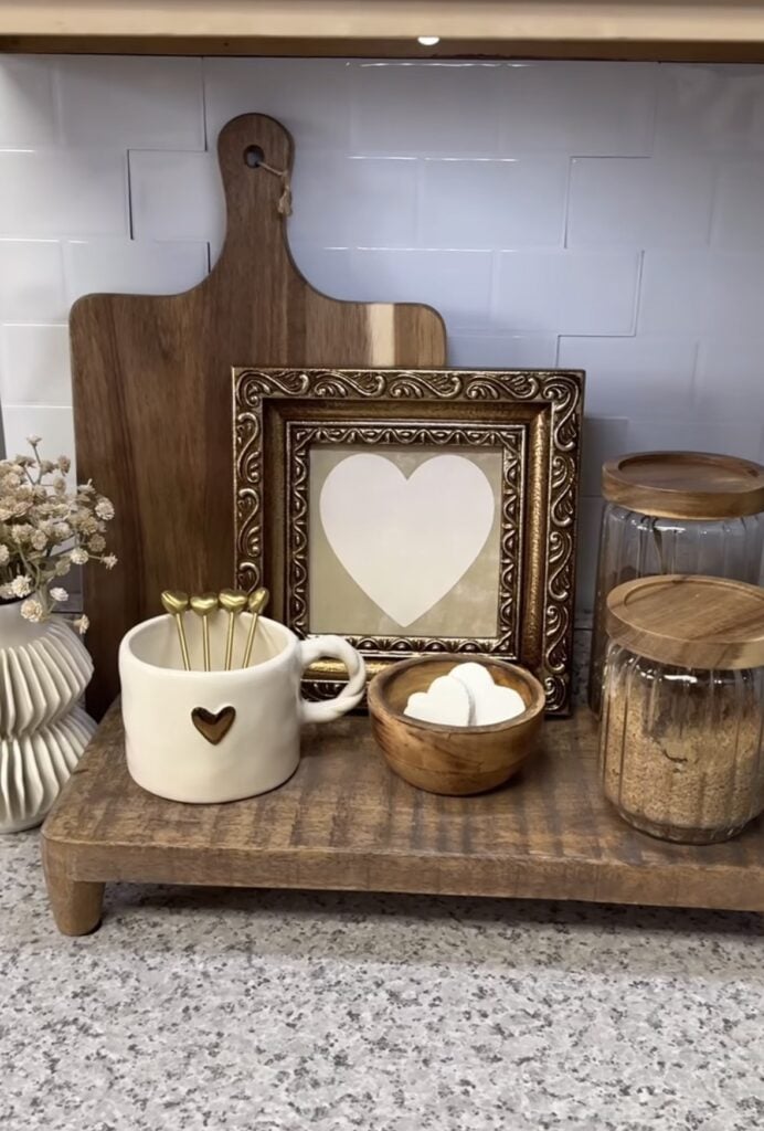 Neutral Valentine’s kitchen counter vignette with a wood cutting board, framed heart art, heart mug with gold heart stir sticks, and a bowl of heart-shaped treats.