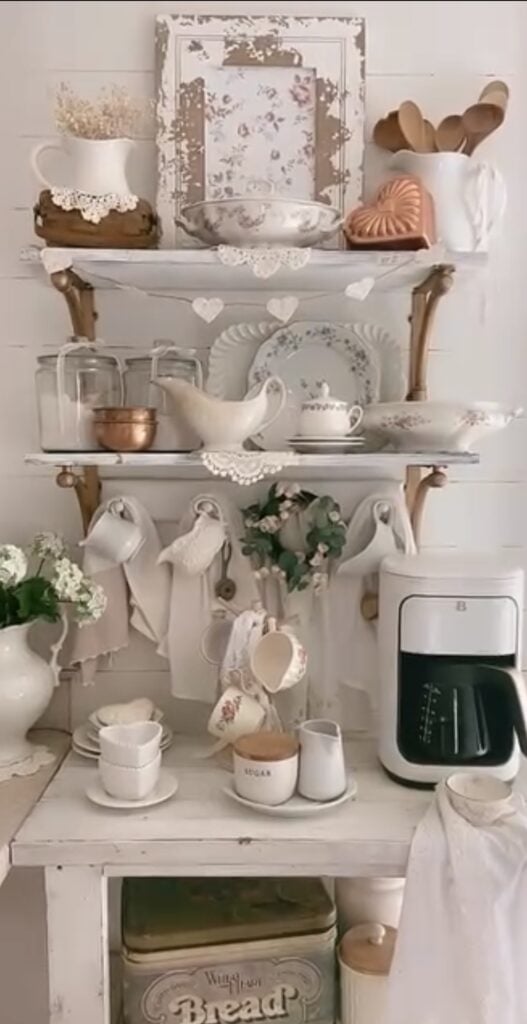 Farmhouse neutral Valentine’s coffee station with open shelves, white dishes, lace accents, heart garland, hanging mugs, and a countertop coffee maker.