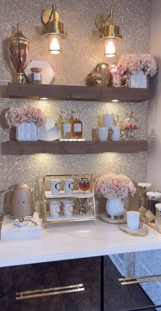 Glam neutral Valentine’s coffee bar with floating shelves, gold heart decor, pink flowers, mugs, and warm under-shelf lighting.