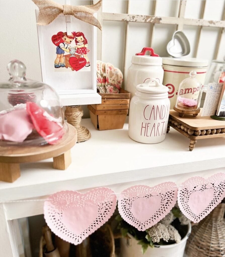 Valentine shelf decor with a “Candy Hearts” canister, vintage jars and tins, a glass cloche, and a pink paper heart garland draped across the front.