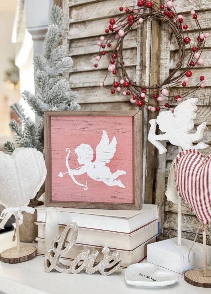 Farmhouse pink Valentine vignette with a pink cupid wall sign, stacked books, a wood “Love” word sign, heart decor on stands, and a berry wreath on rustic shutters.