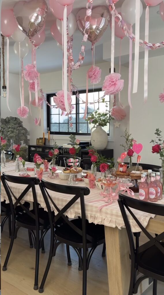 Valentines day dining room table set with pink tablecloth and glass jars along the center with red roses in them with valentines balloons overtop