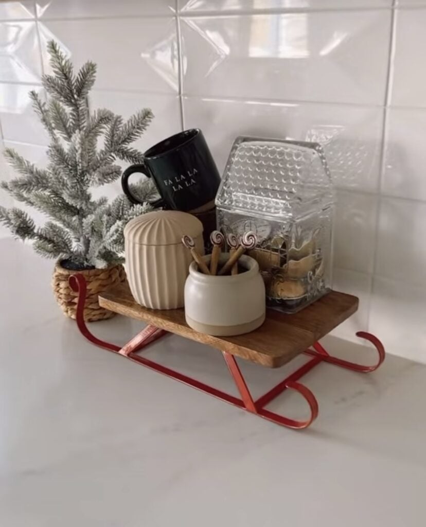 Christmas kitchen vignette on a small sled tray with a bottle brush tree, holiday mug, neutral canisters, and house-shaped cookie jar, featured by slowestuff.com.