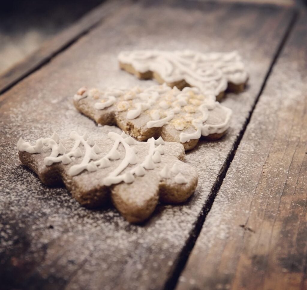 Tree-shaped gingerbread cookies with vanilla icing arranged on a rustic wood board dusted with powdered sugar.”