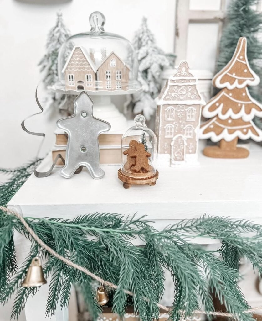 Shelf display with glass cloche gingerbread houses, cookie cutter trees, and garland styled as a gingerbread cloche vignette.