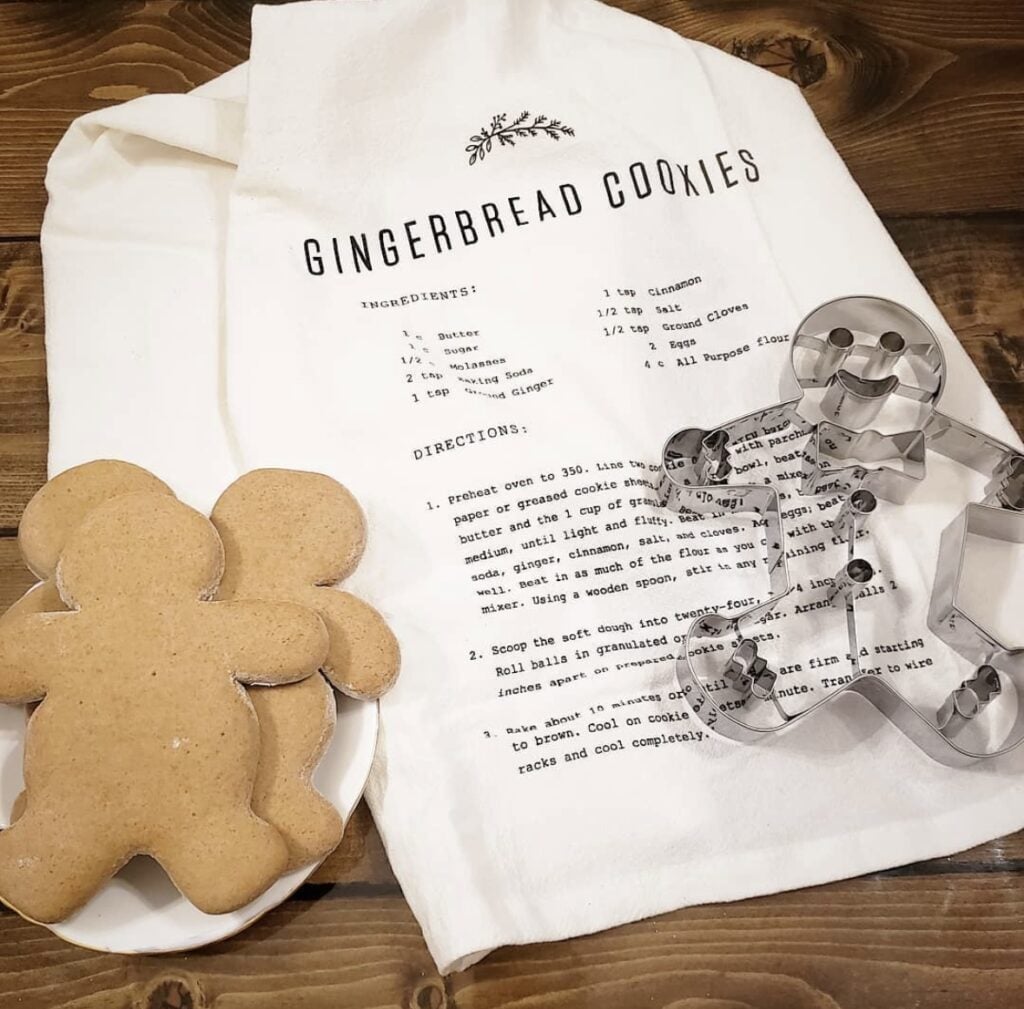 Gingerbread cookie recipe tea towel styled with oversized metal gingerbread cookie cutters and plain gingerbread men on a rustic wood counter.