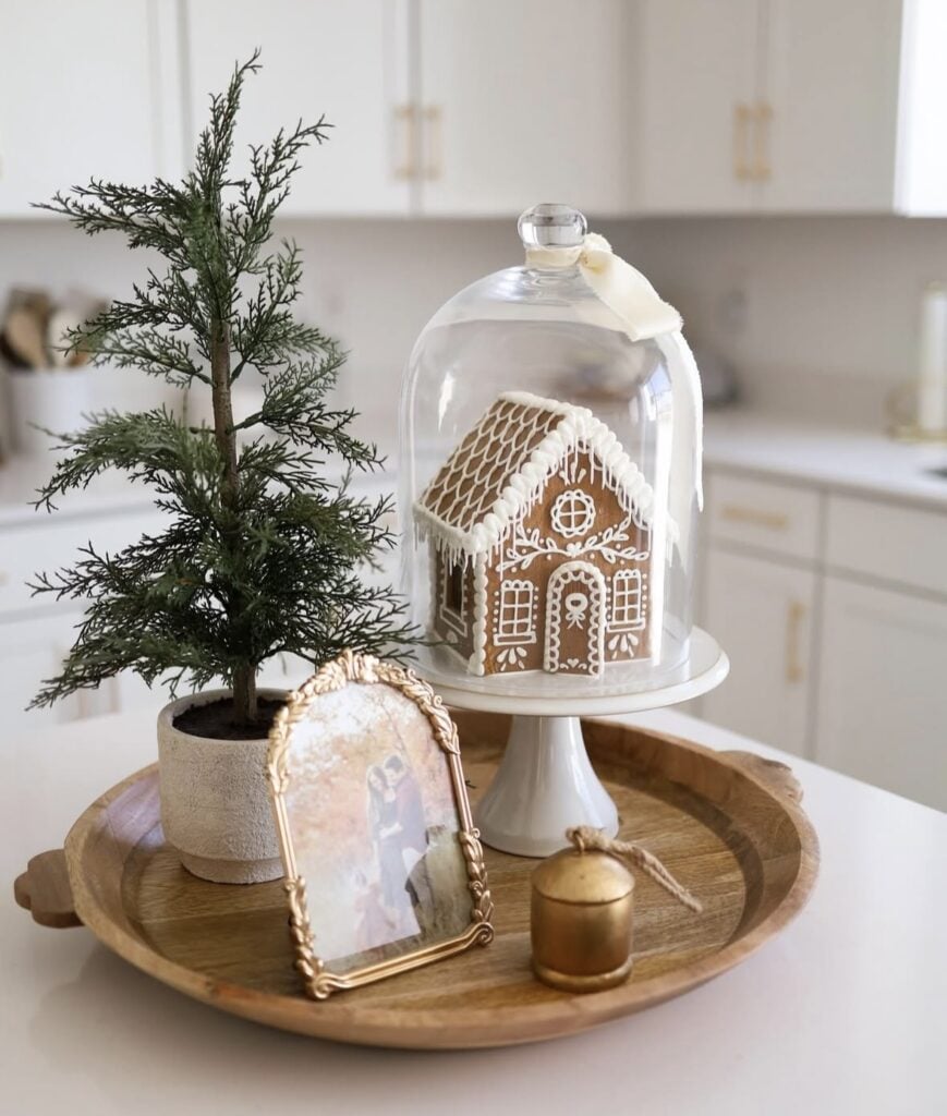 Gingerbread house under a glass cloche on a wooden kitchen tray with a mini Christmas tree and pine sprigs.