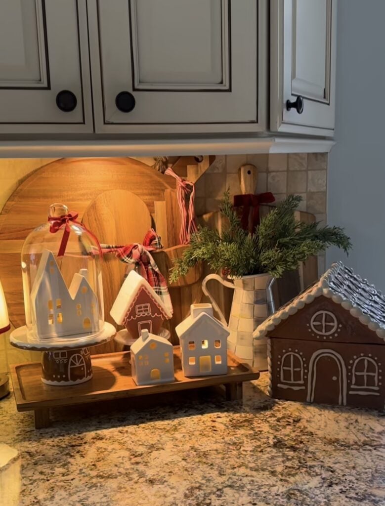 Gingerbread kitchen counter vignette with a small house under a glass cloche, layered cutting boards, and festive Christmas accents, featured by slowestuff.com.