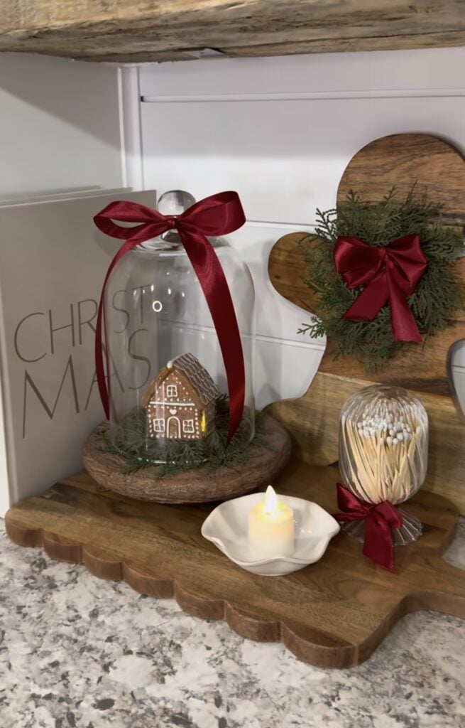 Gingerbread kitchen counter vignette with a glass cloche, miniature house, red bows, cutting board, and festive Christmas decor, featured by slowestuff.com.