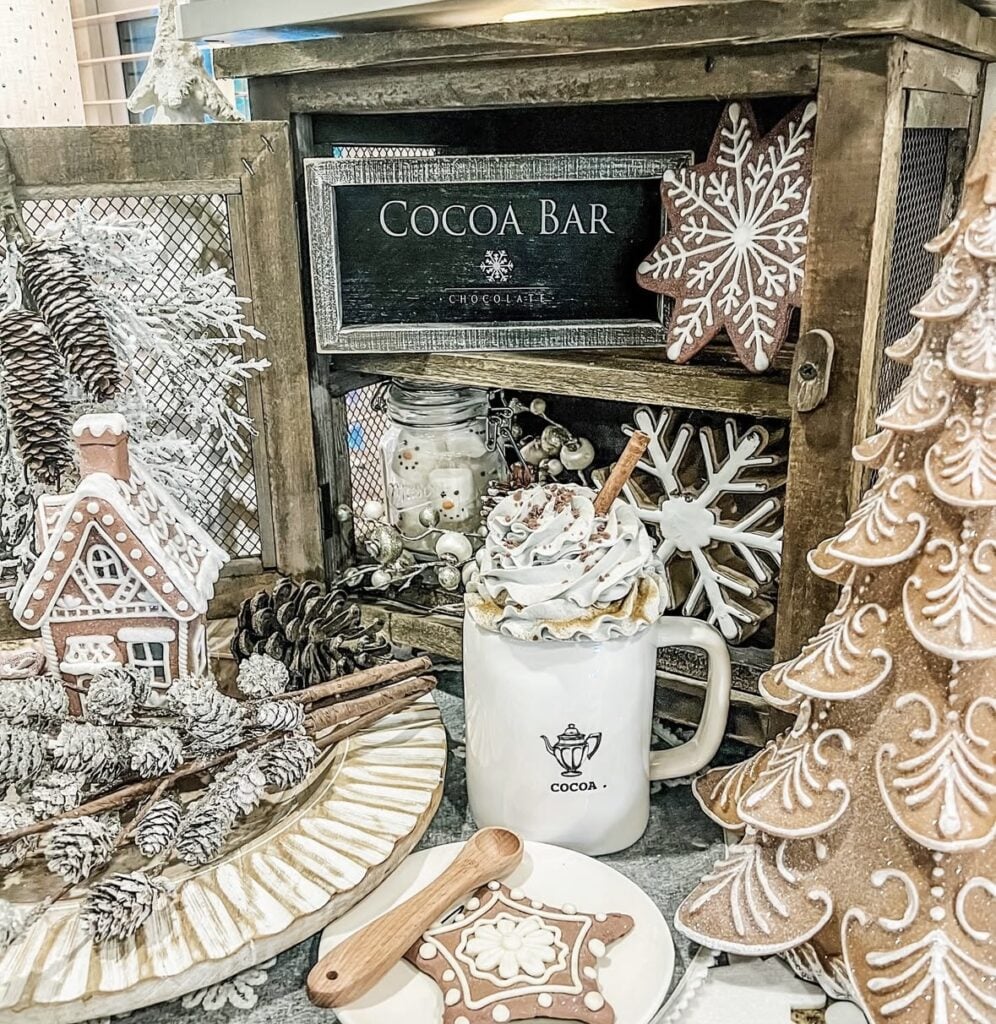 Rustic gingerbread hot cocoa bar with gingerbread houses, snowflake decor, mugs, greenery, and layered Christmas accents, featured by slowestuff.com.