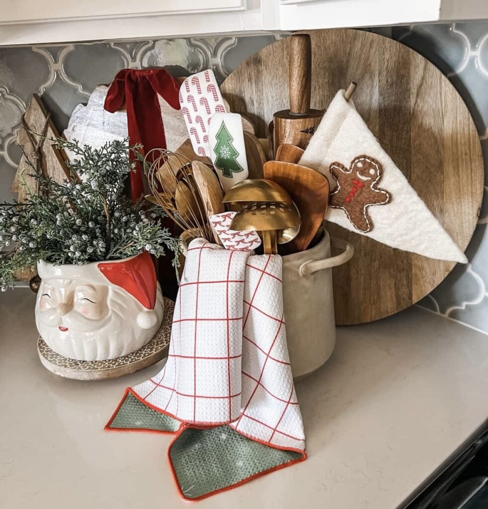 Kitchen utensil crock with spatulas, Santa mug, gingerbread pennant towel, and tiny cookie accents on a bright white counter, featured by slowestuff.com.