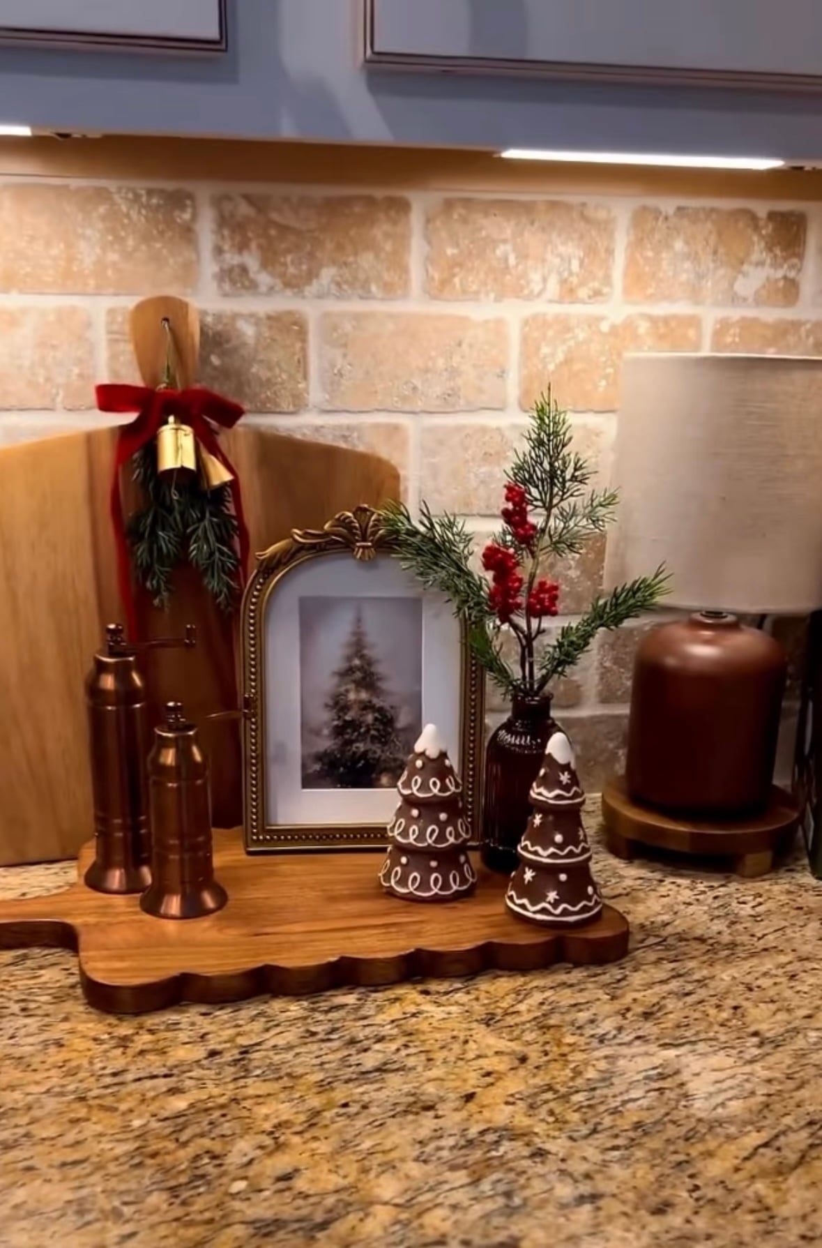 Warm Christmas kitchen backsplash with layered cutting boards, gingerbread houses, small trees, and a brass lamp for cozy evening glow, featured by slowestuff.com.