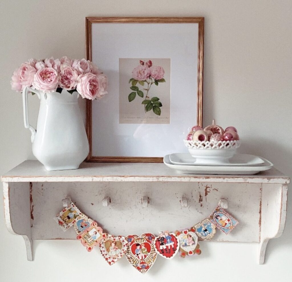 Rustic light pink Valentine’s Day shelf decor with a white pitcher of pink roses, framed floral art, and a heart banner garland.