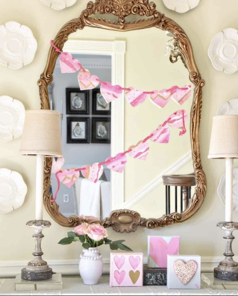 Entry table Valentine’s Day decor with an ornate gold mirror, pink heart banner, lamps, and pink Valentine cards and signs