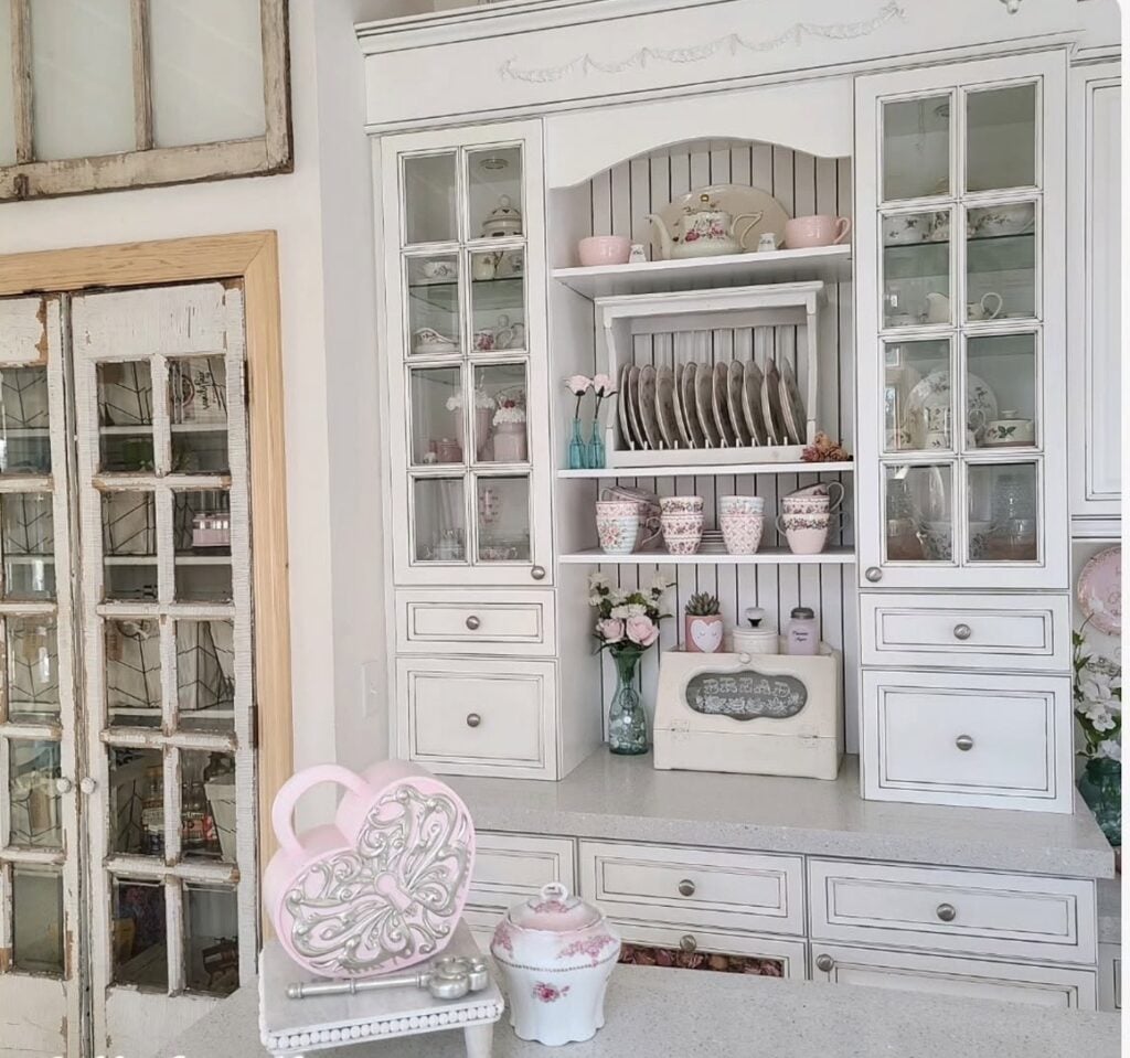 Vintage white hutch styled for Valentine’s Day with pink mugs, floral teacups, stacked dishes, and soft pink accents.