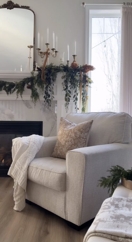 Soft neutral living room with a Christmas fireplace mantel decorated with evergreen garland and brass candlesticks, featured by slowestuff.com.