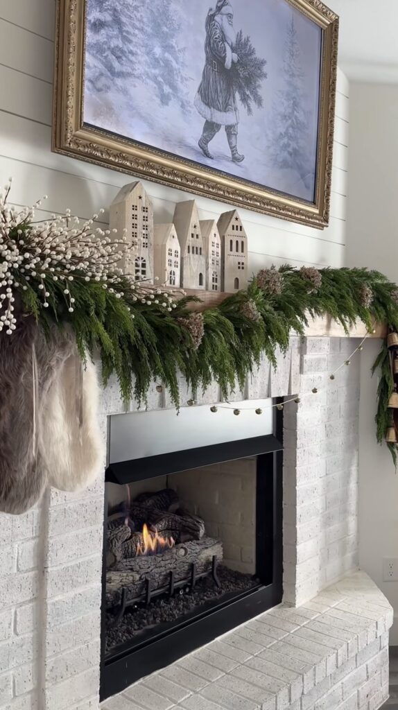Neutral Christmas fireplace mantel decorated with winter Santa artwork, evergreen garland, and small wooden village houses, featured by slowestuff.com.