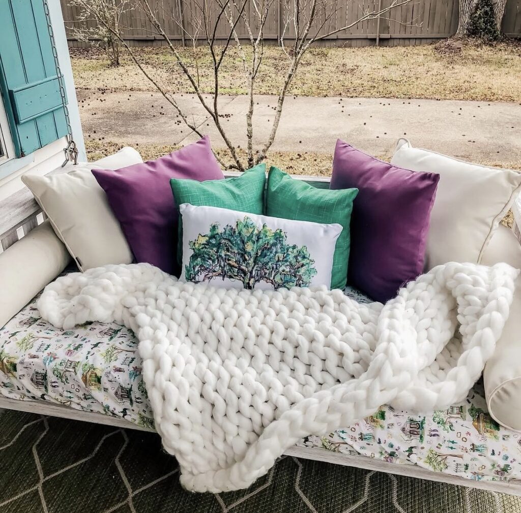 Neutral porch swing styled for Mardi Gras with layered pillows, purple and green accents, and a cozy throw blanket.