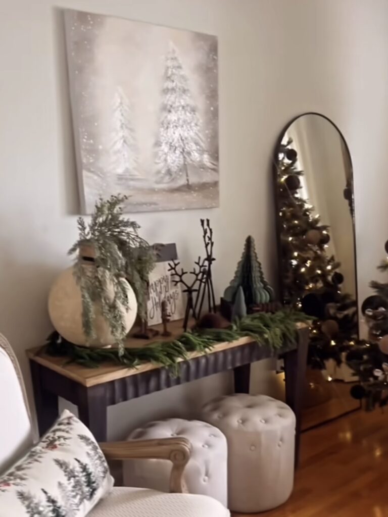 Neutral Christmas side table display with snowy tree artwork, evergreen garland, and cozy upholstered ottomans, featured by slowestuff.com.