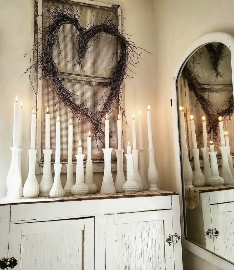 Neutral Valentine vignette with white taper candles in milk glass holders beneath a twig heart wreath on a vintage window screen