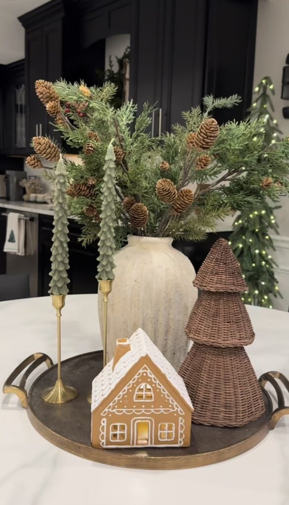 Round tray with neutral gingerbread house, rattan Christmas trees, and an evergreen vase for minimalist Christmas decor.