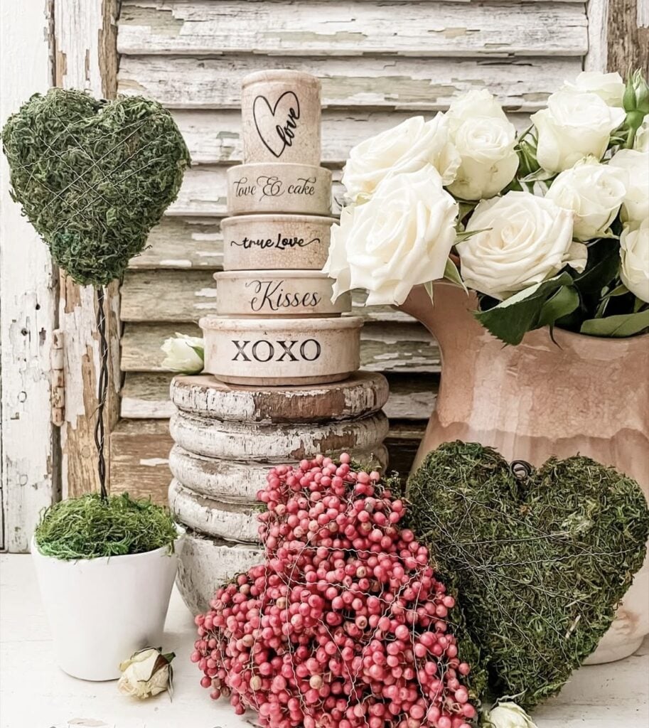 Rustic Valentine’s Day decor with moss heart topiaries, stacked round boxes with love words, white roses, and pink berry hearts.