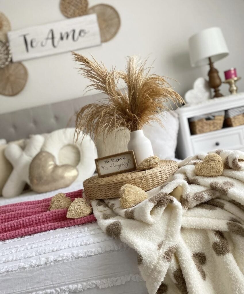 Neutral bedroom Valentine’s Day decor with pampas grass in a white vase, heart pillows, cozy throw blanket, and soft pink bedding accents.