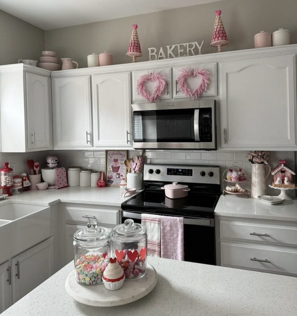 White kitchen Valentine’s Day decor with soft pink accents, heart wreaths on the cabinets, “BAKERY” sign styling, and candy jars on the island.