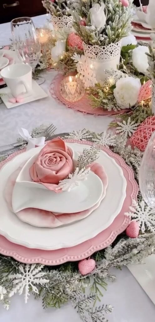 Pink Valentine tablescape with layered blush plates, a rose napkin fold, frosted greenery garland, candlelight, and white floral centerpieces.