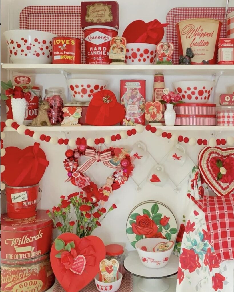 Vintage Valentine kitchen hutch with red and white dishes, candy tins, heart decor, and garland.