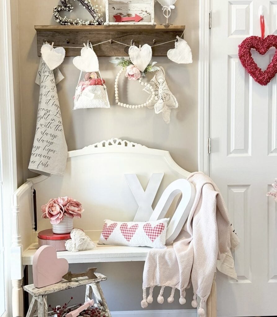 Cozy pink Valentine bench setup with large white “XO” letters, a heart-print pillow, blush throw blanket, and a hanging heart garland on hooks above the bench.