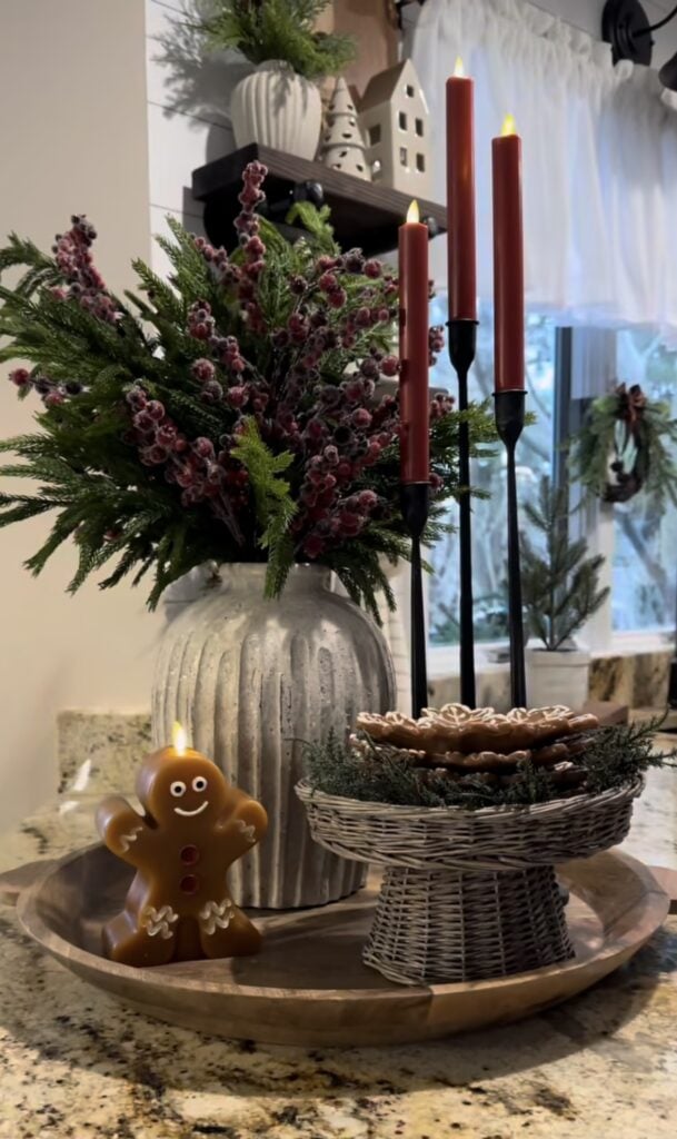 Rectangular tray centerpiece with red taper candle, berry stems, and gingerbread house decor for a Christmas dining table.