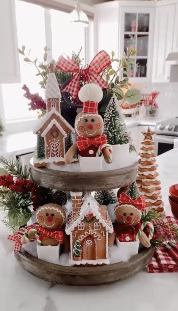 Gingerbread house on a tiered tray with plush gingerbread cookie people and a big red plaid bow for Christmas.