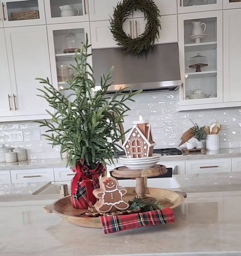 Large gingerbread house and trees on a round tray with red plaid ribbon styled as a Christmas kitchen island centerpiece.