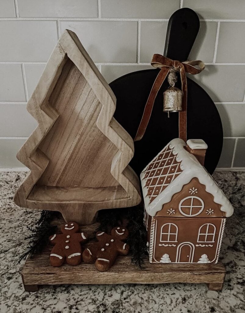 Wooden tray vignette with gingerbread house, wood tree cutting board, and greenery styled for cozy Christmas decor.