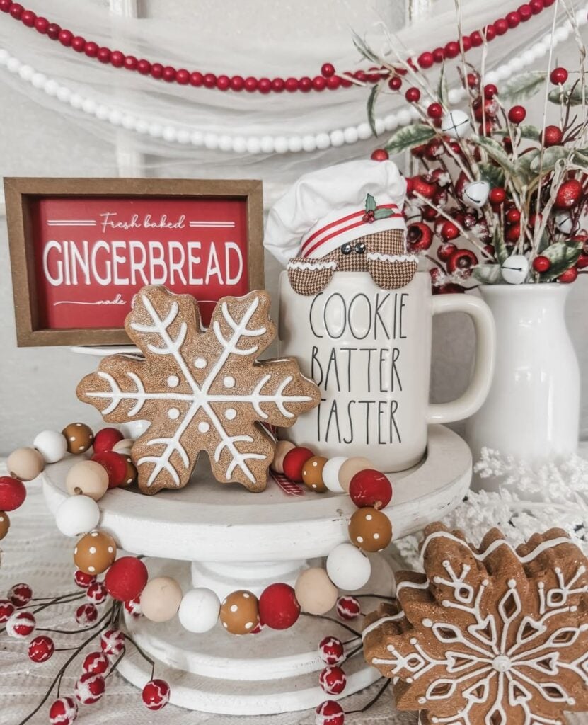 Small pedestal tray with gingerbread house, snowflake cookie, mug, and evergreen sprigs for Christmas kitchen decor.