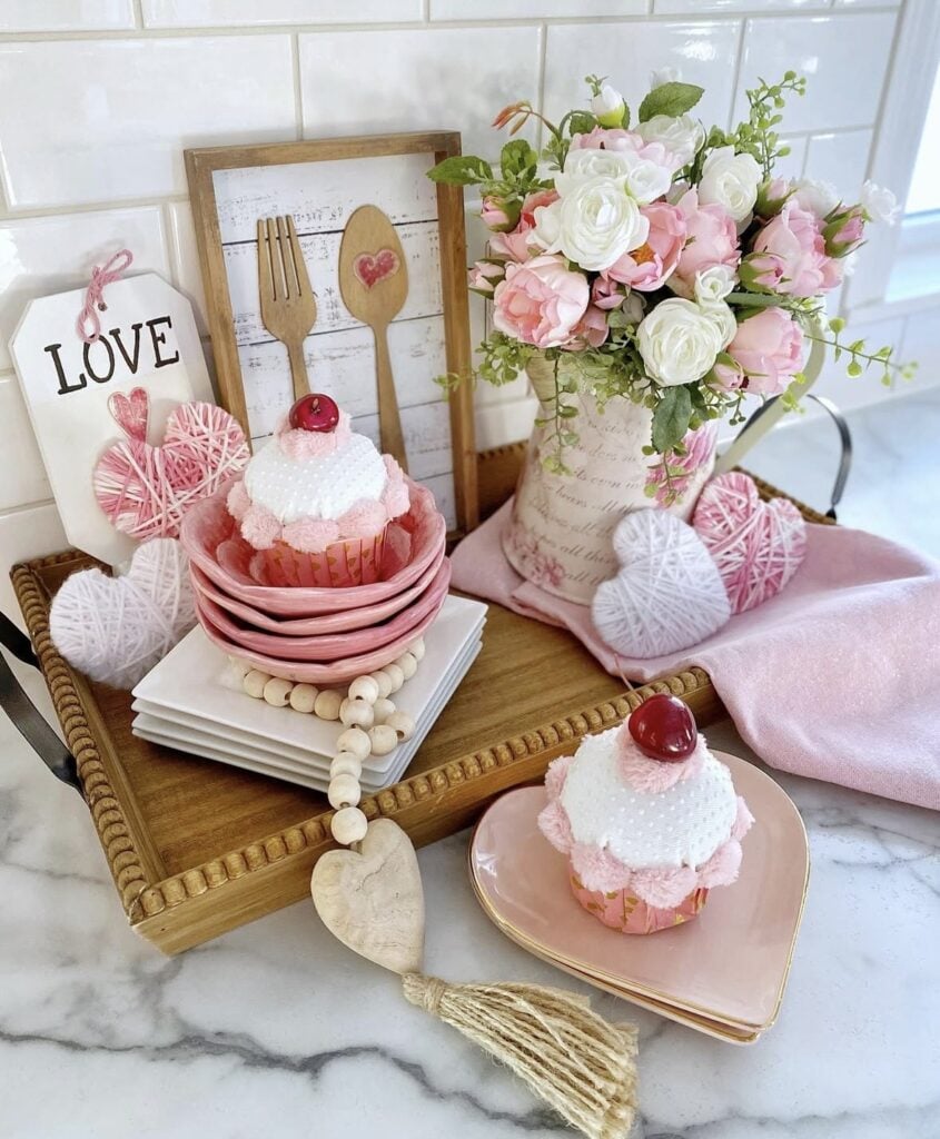 Pink Valentine kitchen tray decor with stacked blush plates, cupcake-style ornaments, yarn-wrapped heart decor, a floral mug arrangement, and a pink cloth runner on marble