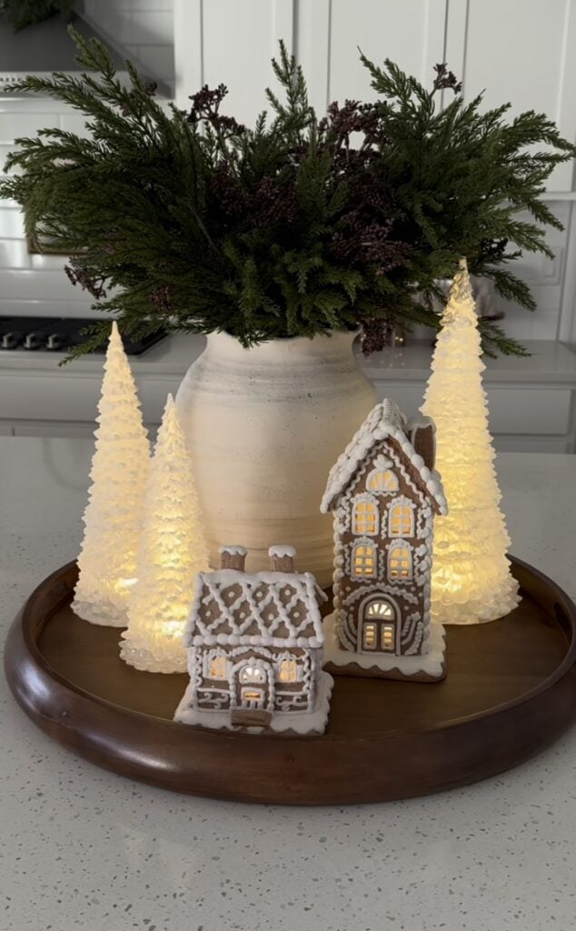 Winter white gingerbread houses on a tray with lit trees and greenery in a glass vase for soft neutral Christmas decor.