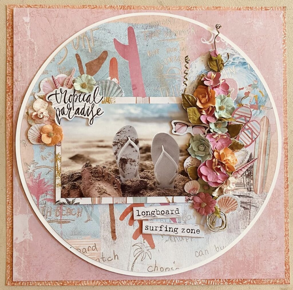Beach scrapbook layout with flip flop photo, “tropical paradise” title, layered patterned papers, and a floral embellishment cluster with seashells and sunglasses.