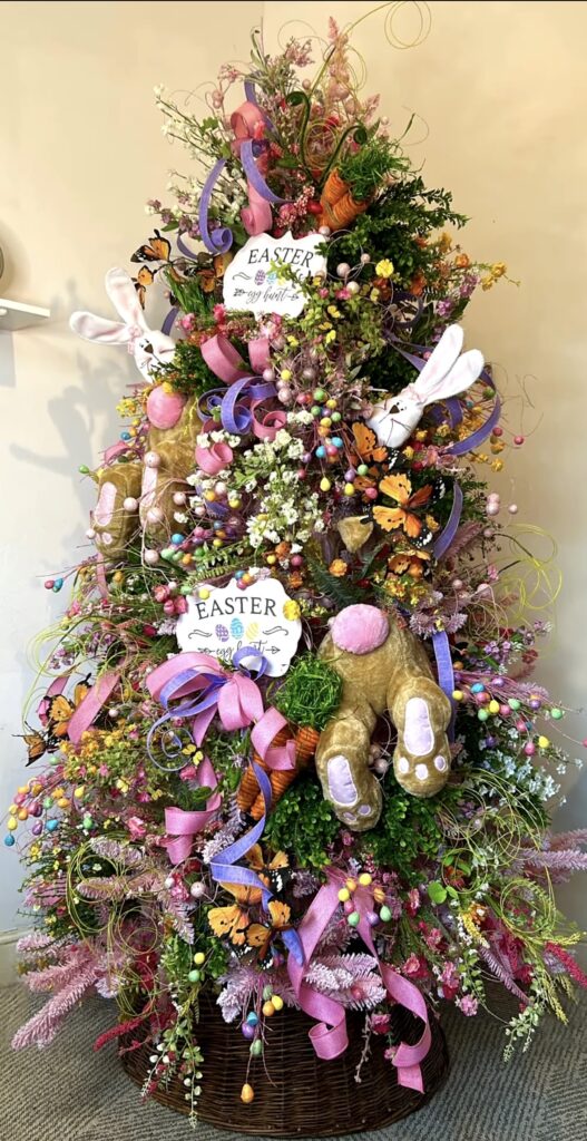 Over-the-top Easter tree filled with bunny legs, pastel ribbons, spring flowers, colorful egg picks, and “Easter egg hunt” signs.