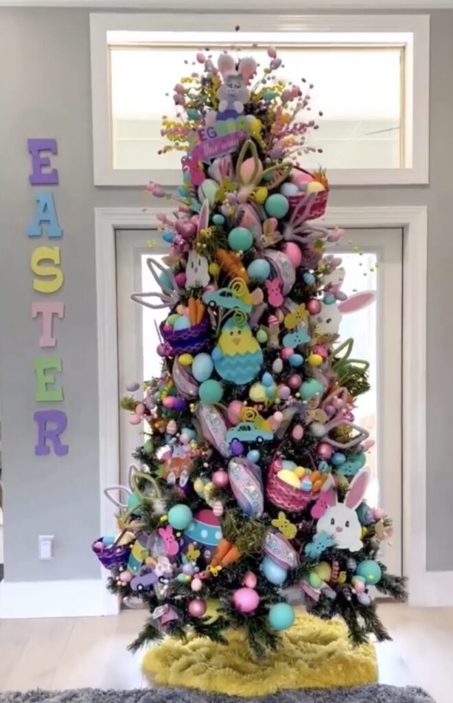 Bright and colorful Easter tree packed with pastel eggs, bunny picks, baskets, and spring decorations, displayed beside an “EASTER” letter sign on the wall.
