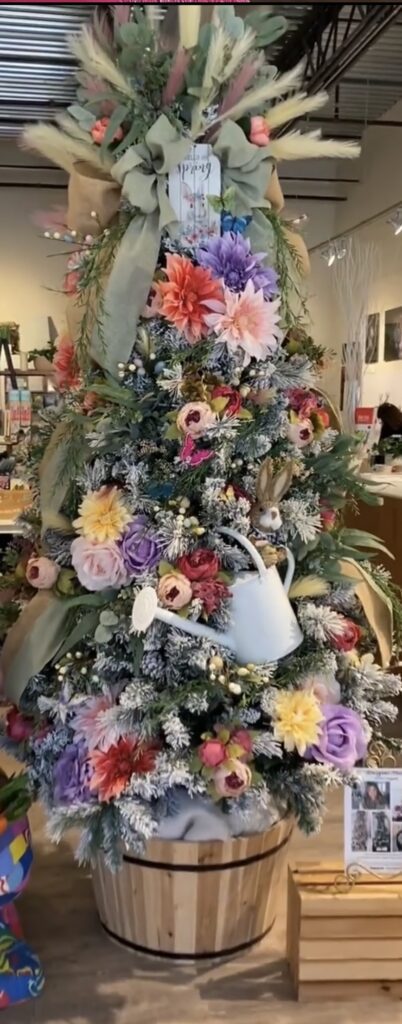 Rustic spring Easter tree decorated with colorful flowers, bunny ornaments, greenery, and a white watering can accent in a wooden planter base.