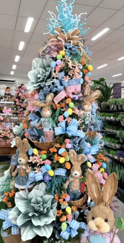 Large Easter tree display with bunny figures, colorful egg ornaments, blue ribbons, spring florals, and pastel greenery