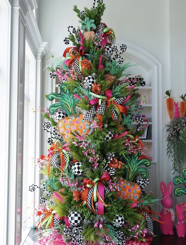 Whimsical Easter tree decorated with oversized carrot ornaments, black and white checkered ribbon, bright pink flowers, and green foliage.