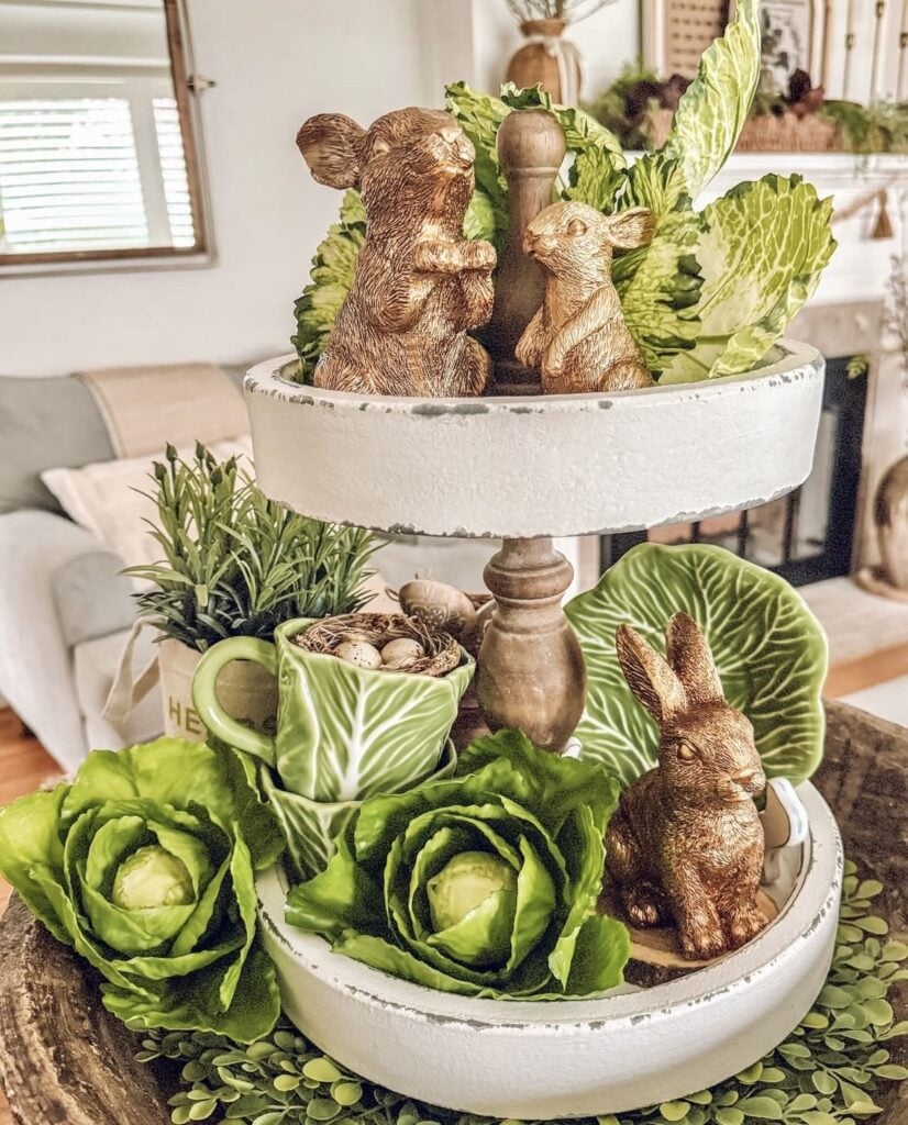 Easter tiered tray decorated with gold bunny figurines, faux cabbage leaves, green ceramic mug, and neutral farmhouse decor on a white two tier tray.