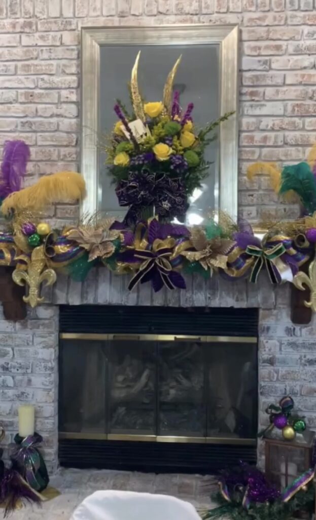 Mardi Gras mantel decor on a white brick fireplace with purple, green, and gold garland, fleur-de-lis accents, feather sprays, and a floral centerpiece.
