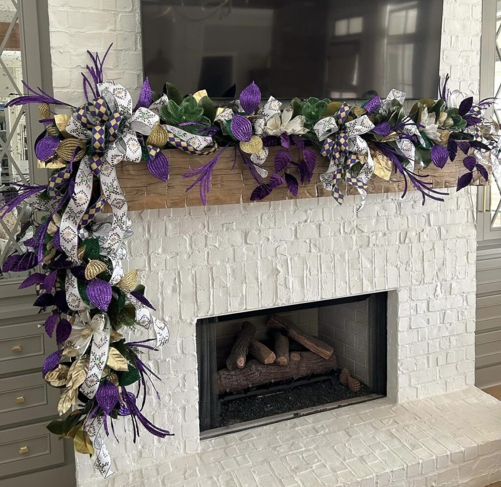 White brick fireplace mantel decorated for Mardi Gras with a lush garland in purple, green, and gold, featuring large bows, metallic leaves, and ribbon tails cascading down the left side.