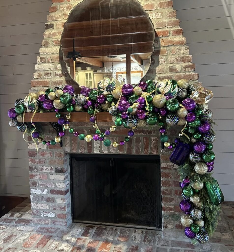Brick fireplace with Mardi Gras ornament garland packed with purple, green, and gold baubles, glitter ornaments, and bead strands draped across the mantel and down the right side.
