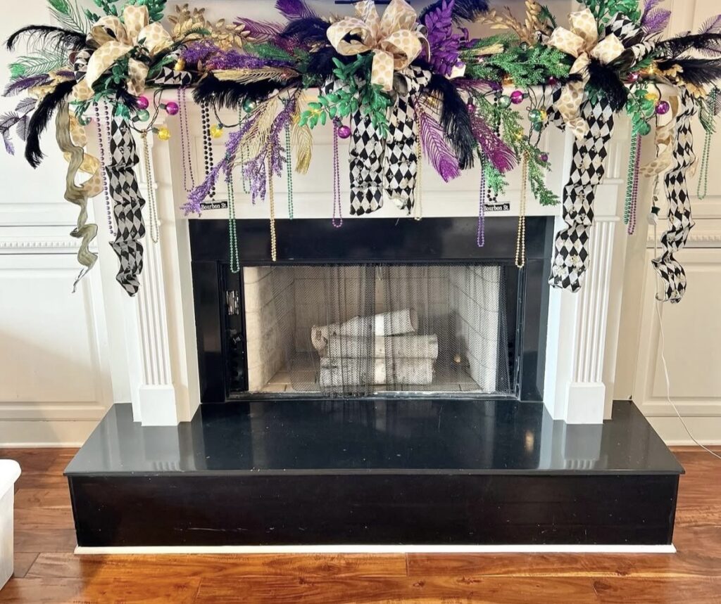 White fireplace mantel with dramatic Mardi Gras swag using feather sprays, greenery, black-and-white harlequin ribbon, gold bows, and hanging bead strands across the top.