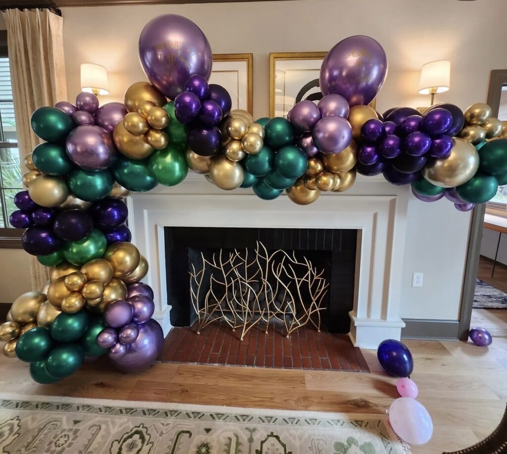 Mardi Gras balloon garland framing a fireplace in metallic purple, green, and gold balloons clustered thickly across the mantel and down the left side.